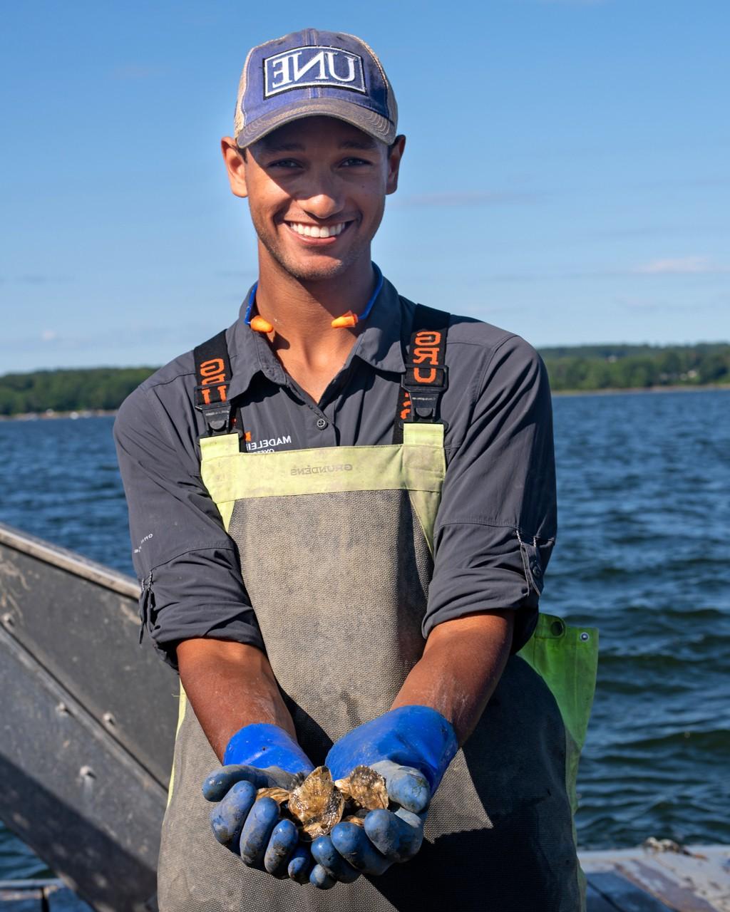 A student wearing a U N E baseball hat and waders shows off several oysters in their hands