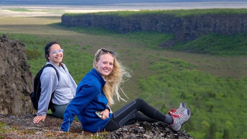 Two students sitting on the edge of a cliff overlooking cliffs and the ocean in Iceland