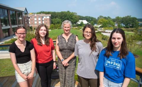 Four students who have received the Millennium Fellowship pose with Alethea Cariddi, associate director of sustainability at UNE, on a campus deck. The campus quad and commons are visible at back.